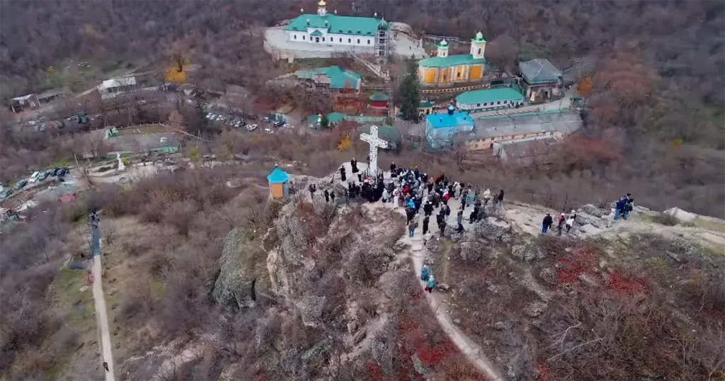 Pilgrims at the base of the Great Cross on Grimidon Rock, overlooking the blue-domed Holy Trinity Monastery in the canyon below.