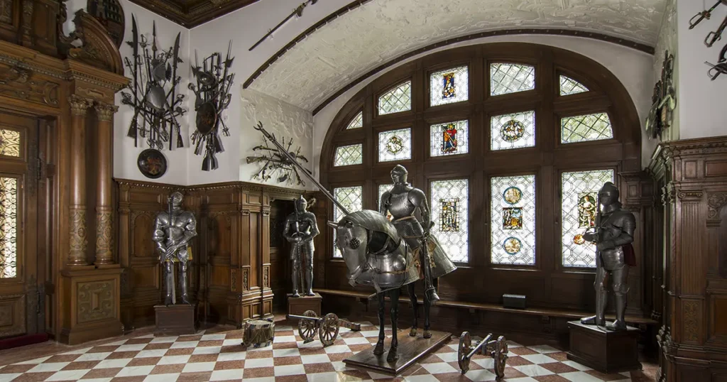Interior view of the Arms Hall at Peleș Castle featuring 16th-century German armor, Eastern weapons, carved wooden walls, and royal military displays.