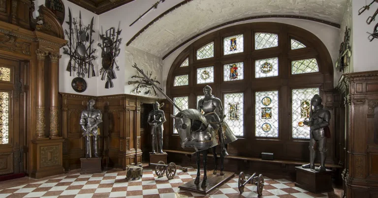 Interior view of the Arms Hall at Peleș Castle featuring 16th-century German armor, Eastern weapons, carved wooden walls, and royal military displays.
