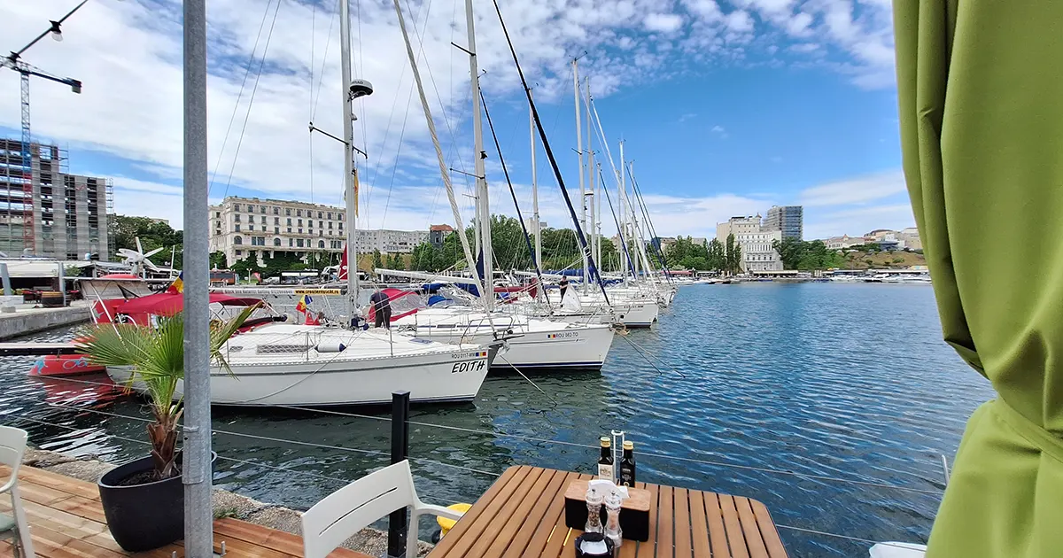Modern yachts and sailboats anchored in the docks of Tomis Marina in Constanța, with waterfront restaurants and terraces lining the harbor under a clear sky.